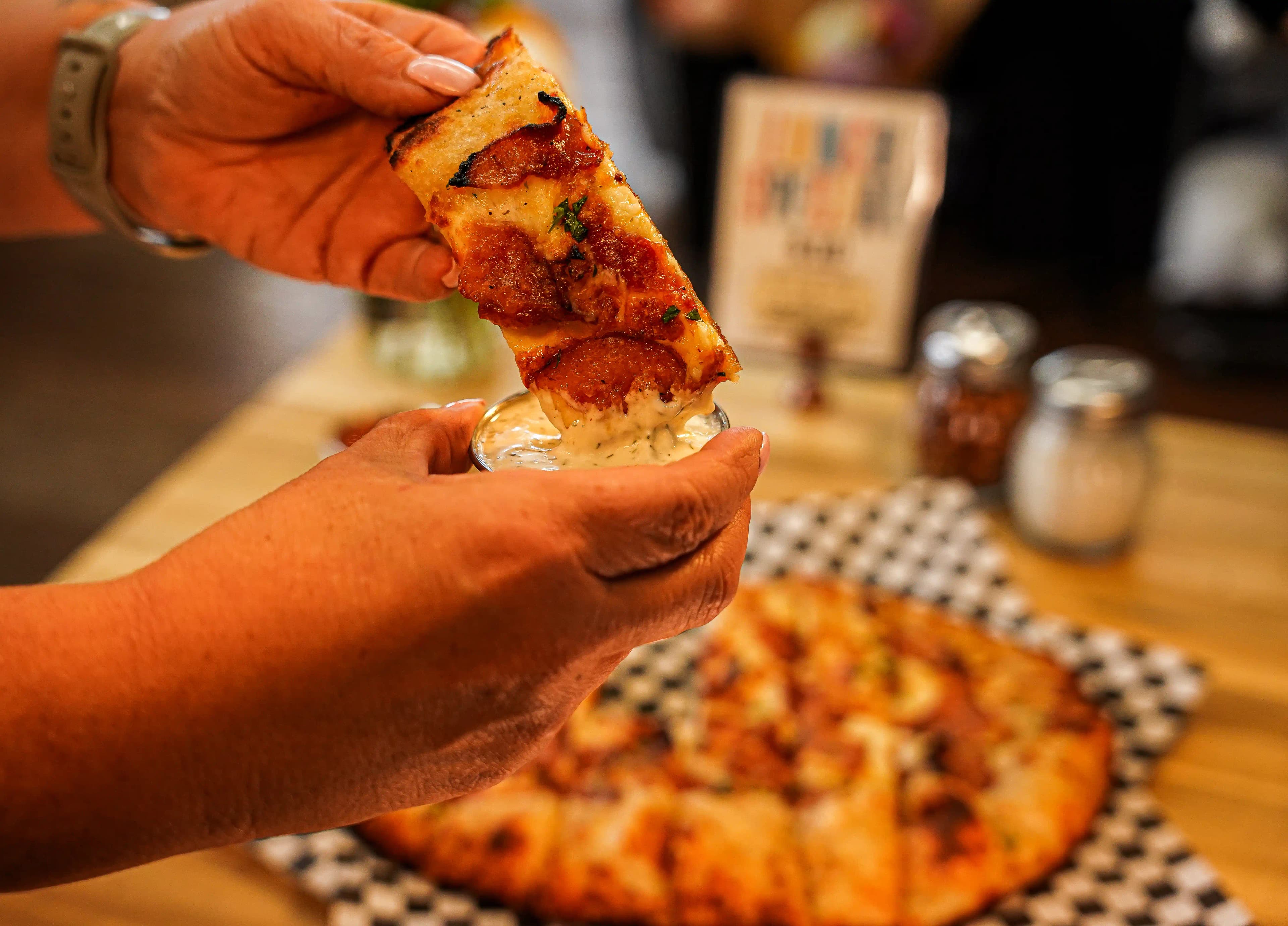 Person dipping a slice of pepperoni pizza into a cup of ranch dressing, with a whole pizza on a checkered tray in the background.
