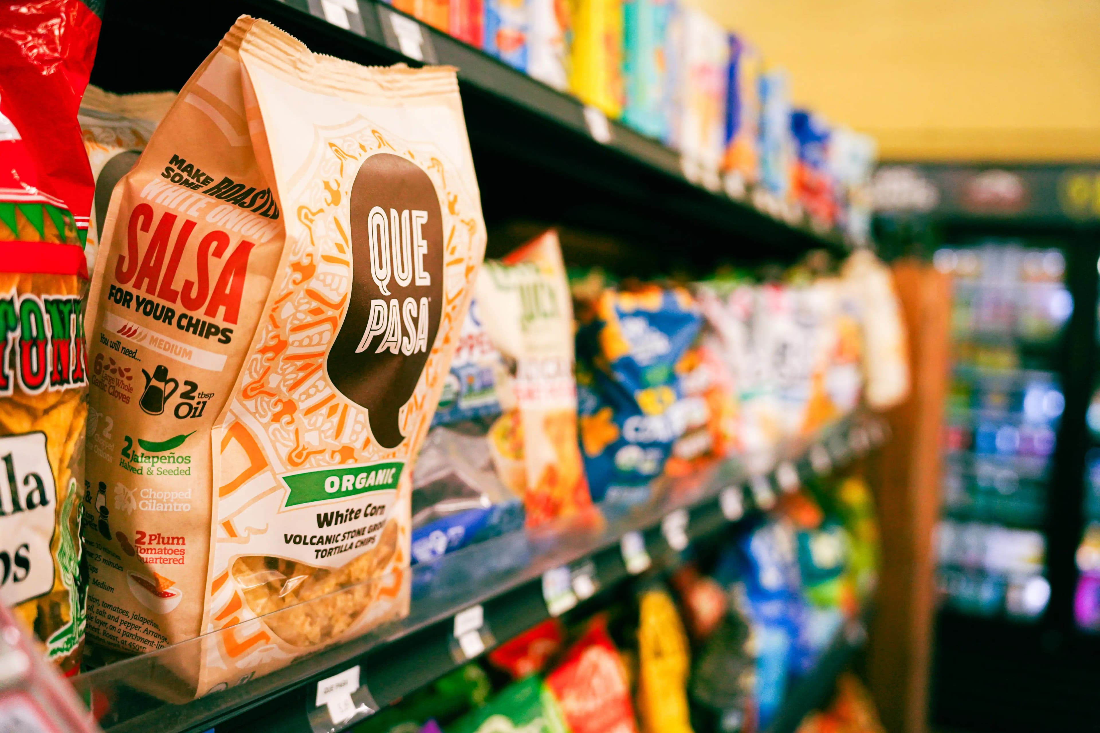 Bags of organic Que Pasa white corn tortilla chips on a grocery store shelf among assorted snack brands.
