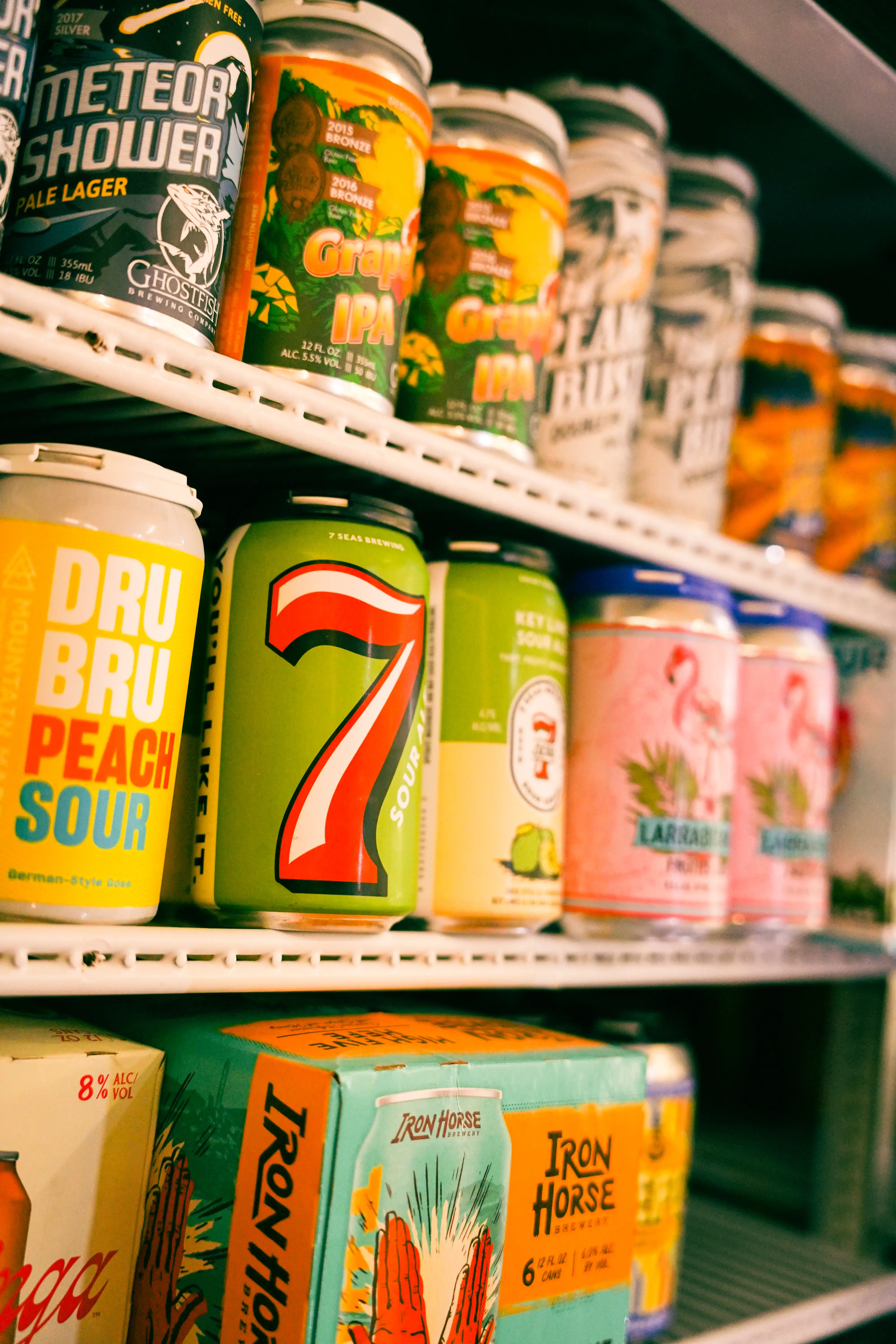 Assorted craft beer and cider cans on a refrigerated store shelf, including 7 Seas, Dru Bru, Iron Horse, and Ghostfish Brewing labels.