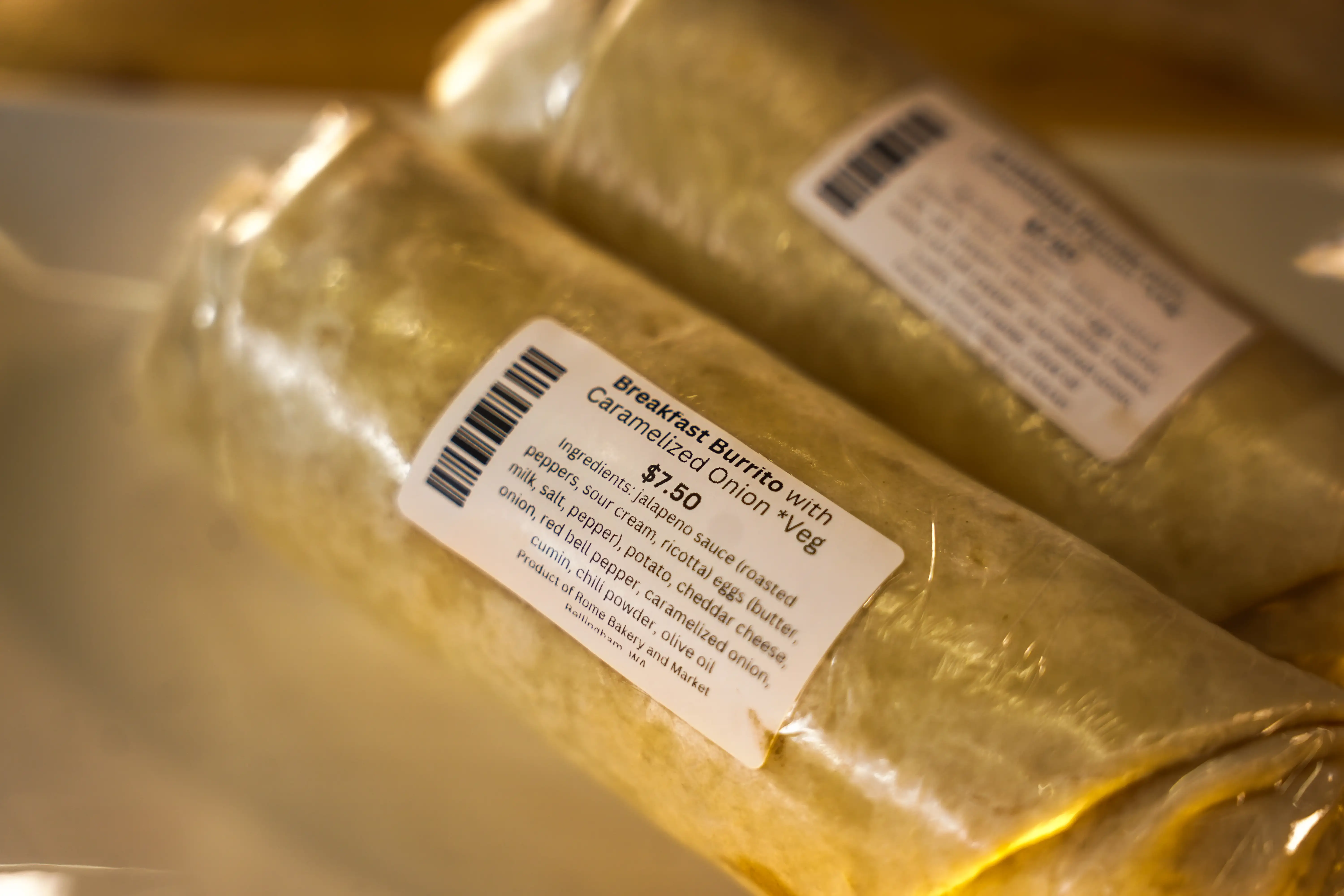 Wrapped breakfast burritos labeled 'Breakfast Burrito with Caramelized Onion + Veg $7.50' displayed on a deli counter.