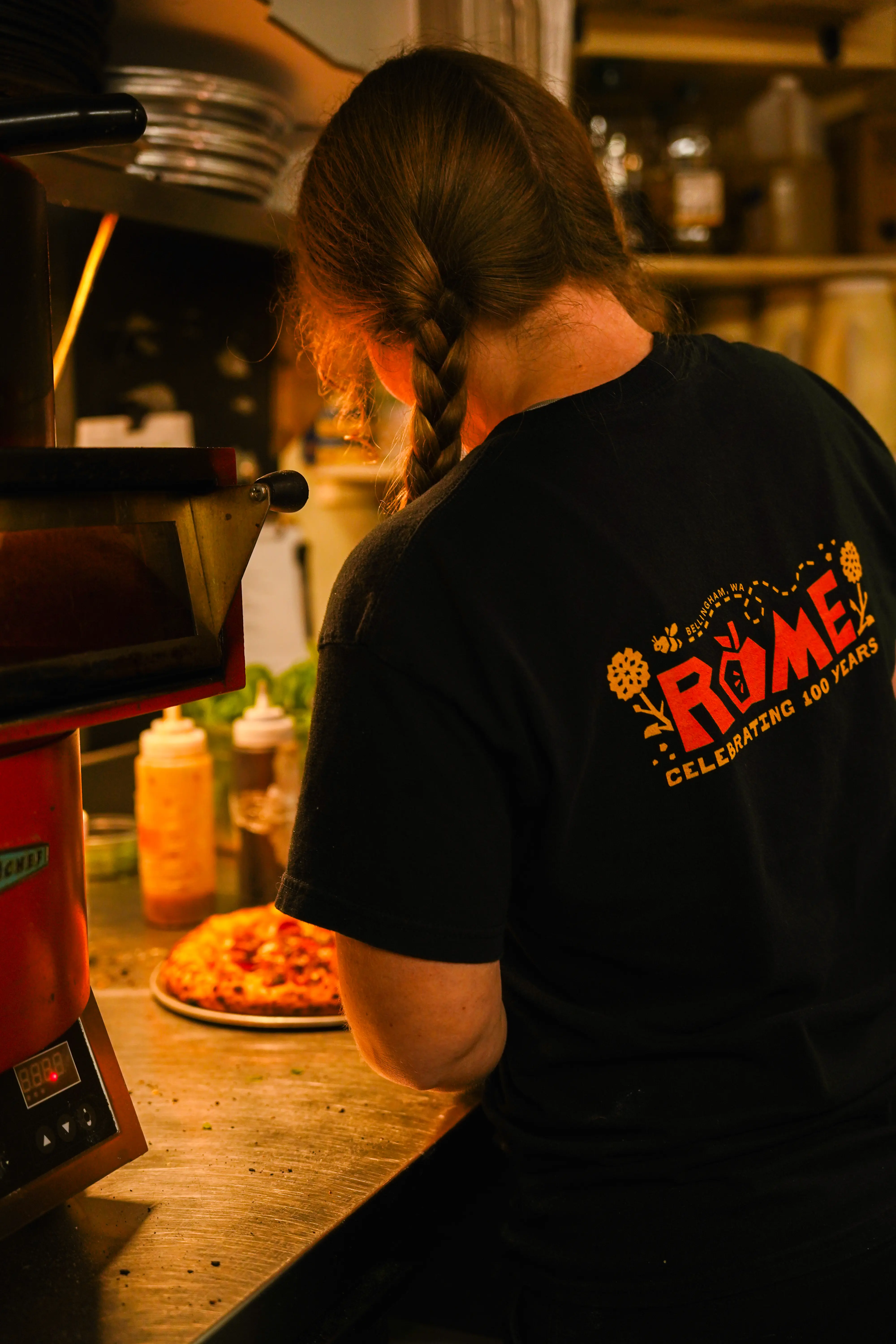 Back of a female employee wearing a black shirt with a red Rome Bakery & Market logo, slicing a pizza.