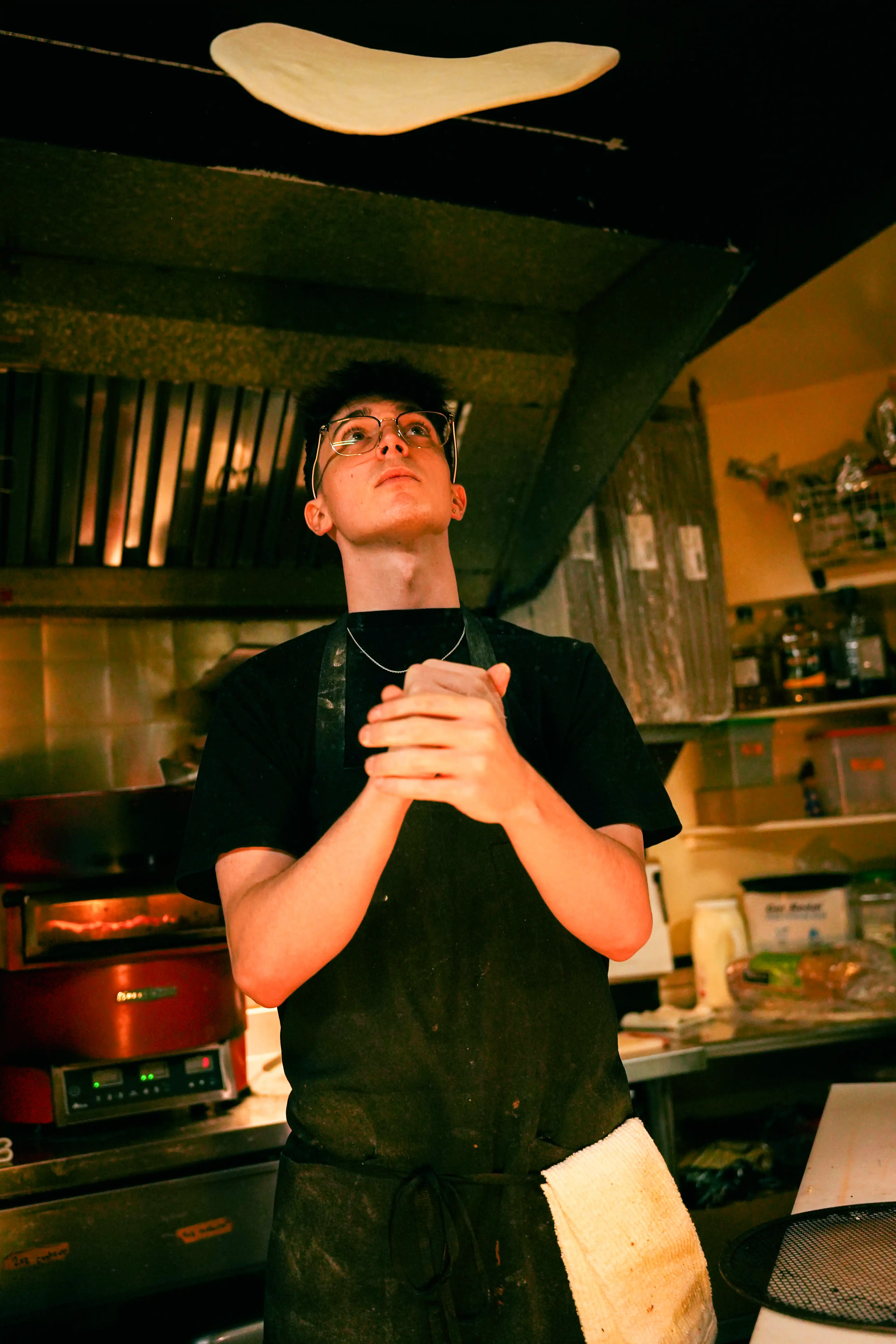 Male employee wearing a black apron tossing pizza dough in the air.