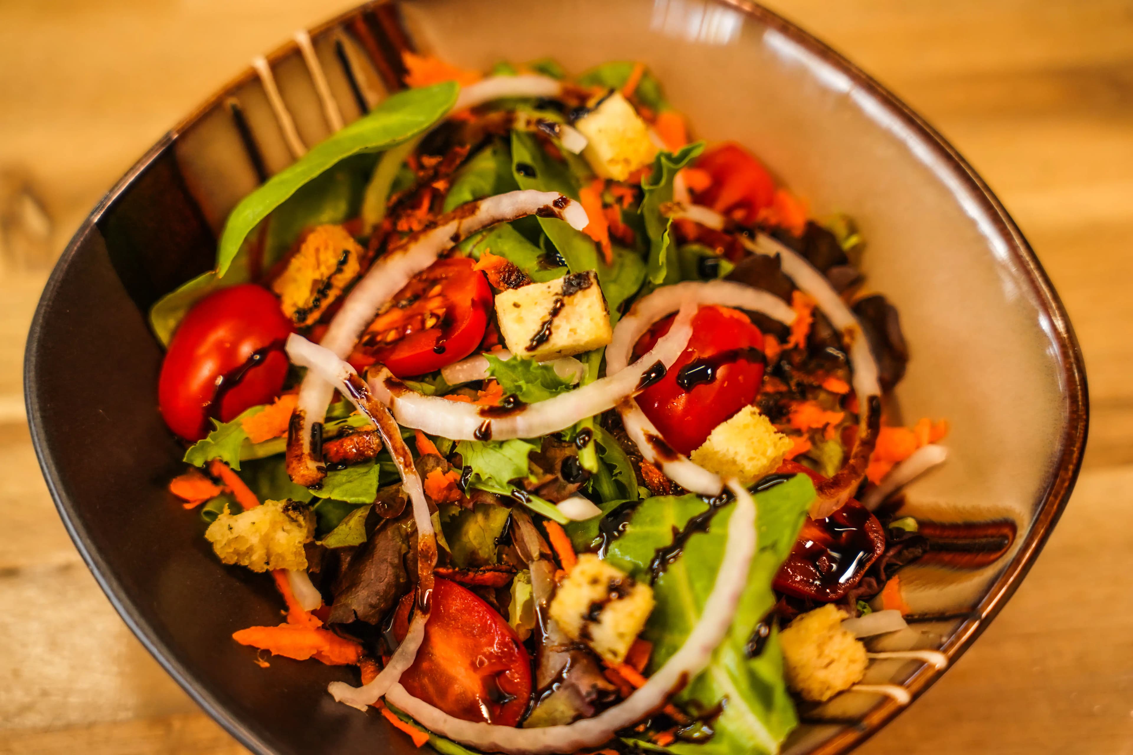 Fresh mixed green salad with cherry tomatoes, croutons, shredded carrots, and sliced onions drizzled with balsamic glaze in a bowl.
