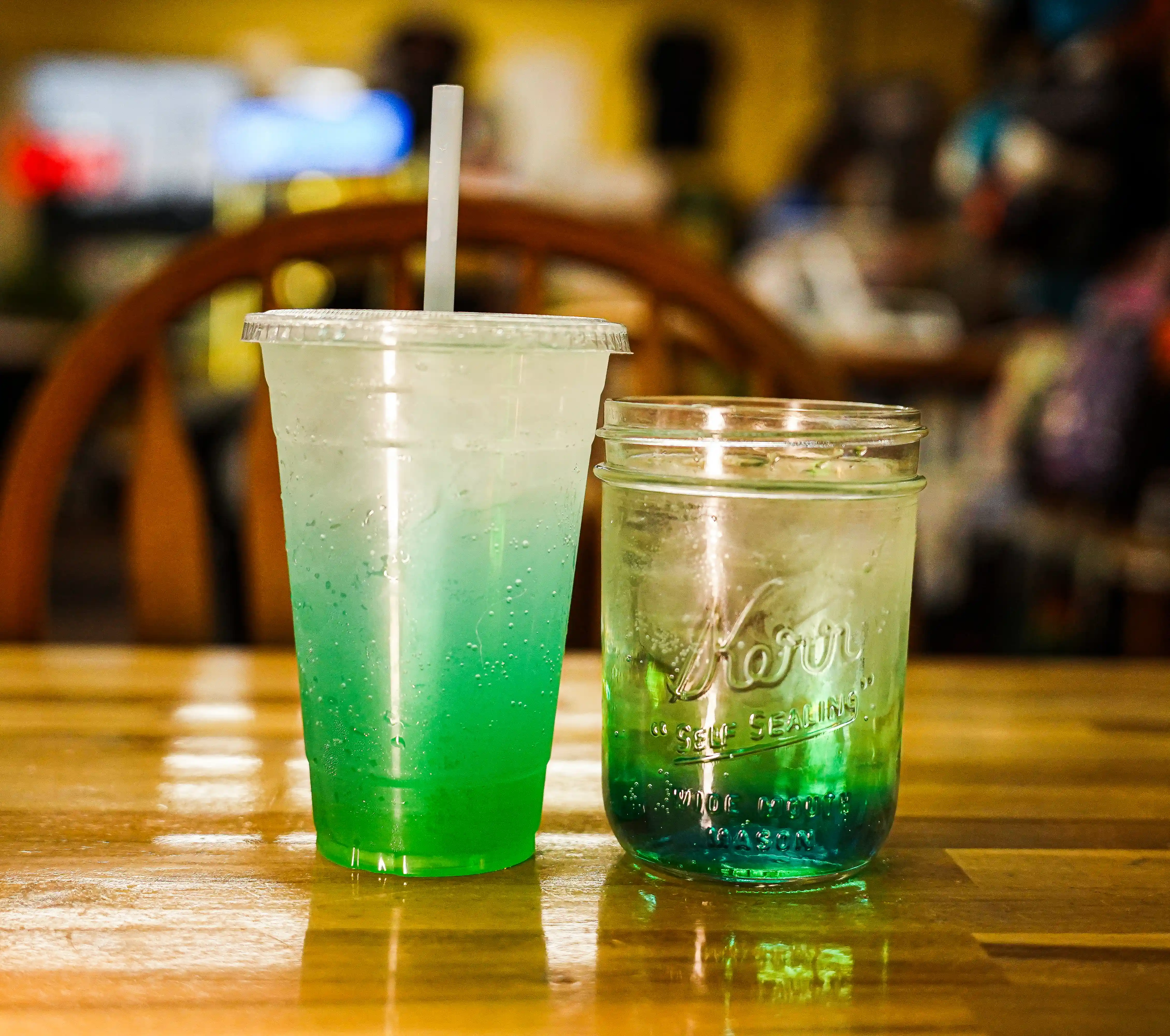 Two green and blue layered sodas on a wooden table, one in a plastic cup with a straw and one in a mason jar.