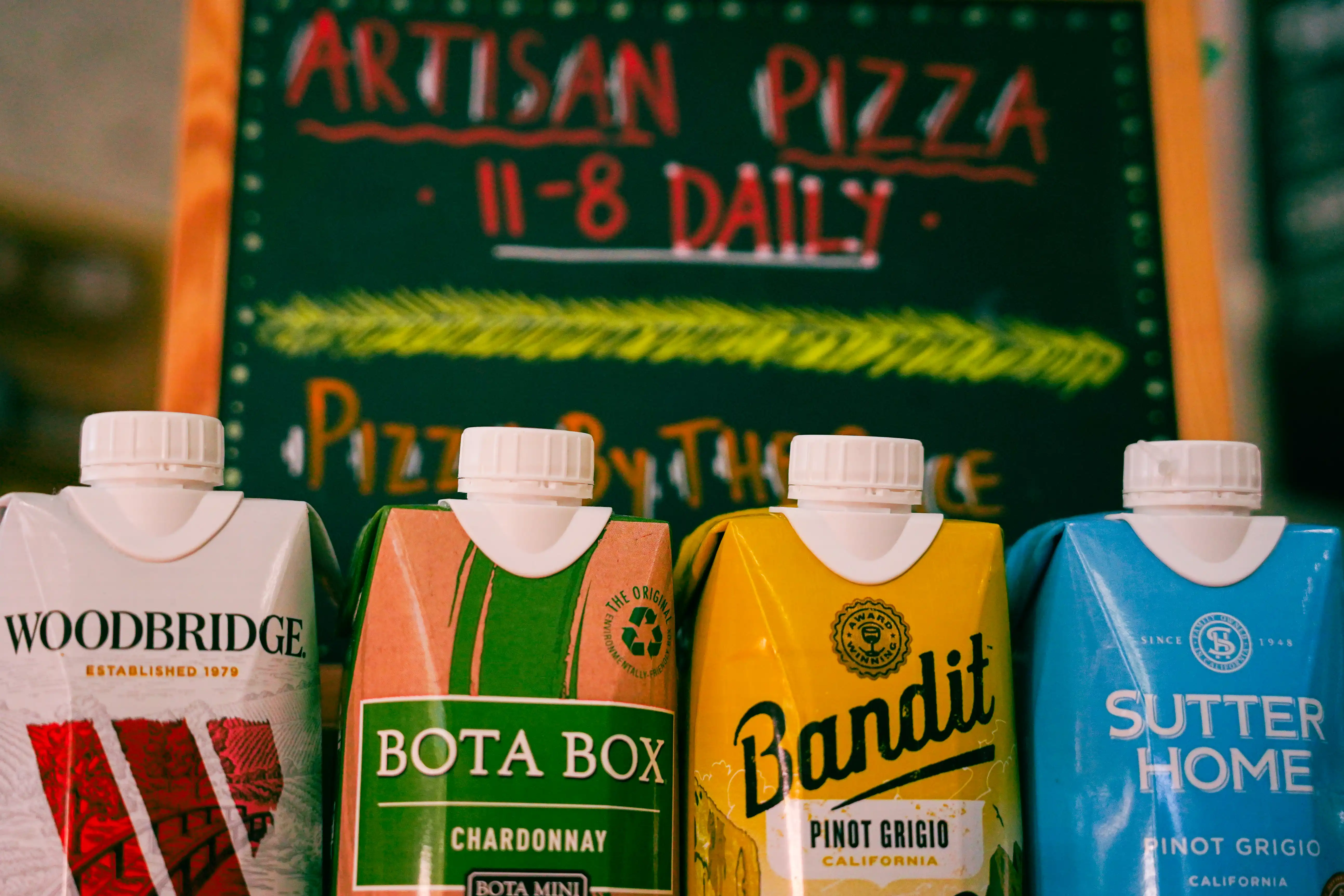 Four boxed wine cartons lined up in front of a chalkboard sign reading 'Artisan Pizza 11–8 Daily,' including Woodbridge, Bota Box, Bandit, and Sutter Home.